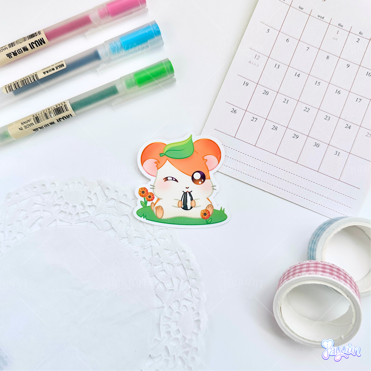 A sticker of Hamtaro from the cartoon Hamtaro sitting on a patch of grass eating a sunflower seed, photographed on a white desk background.