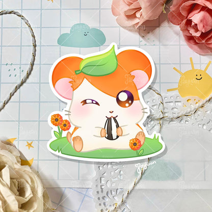 A sticker of Hamtaro from the cartoon Hamtaro sitting on a patch of grass eating a sunflower seed, shown on a decorative paper background.