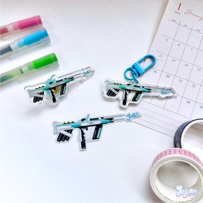 Teal Arc-inspired rifle acrylic pin, acrylic charm, and sticker photographed on a white desk background.