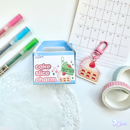 Cake slice charm blind box photographed alongside a strawberry cake slice acrylic keychain on a white desk background,