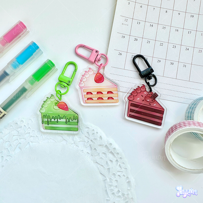 A matcha cake slice acrylic keychain, strawberry cake slice acrylic keychain, and chocolate cake slice acrylic keychain photographed on a white desk background.