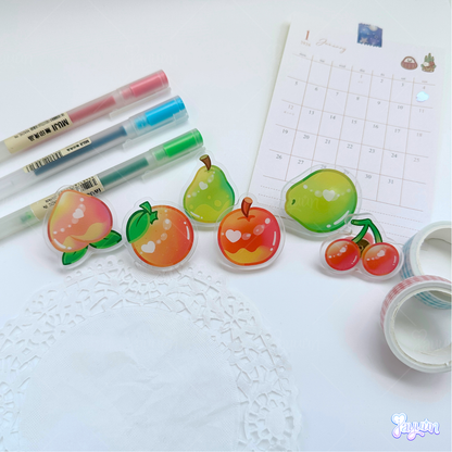 A peach, orange, pear, apple, coconut, and apple acrylic pins photographed on a white desk background.
