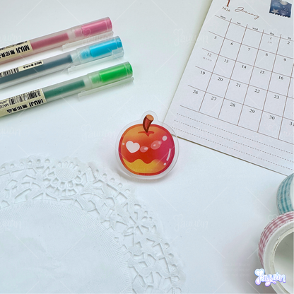 A red apple acrylic pin photographed on a white desk background.