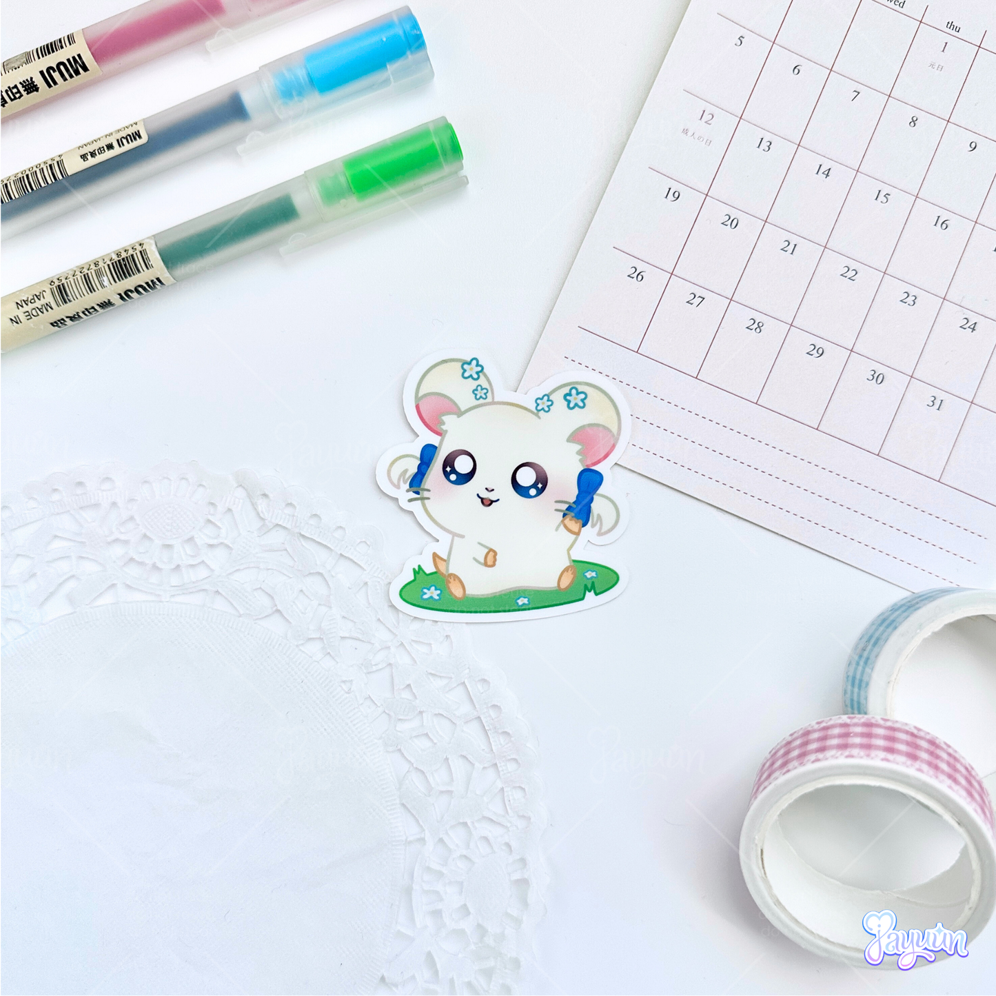 A sticker of Bijou from the cartoon Hamtaro sitting on a patch of grass, photographed on a white desk background.