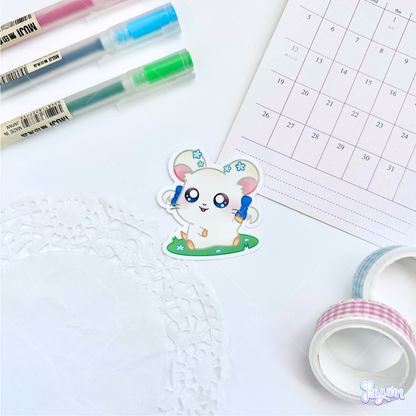 A sticker of Bijou from the cartoon Hamtaro sitting on a patch of grass, photographed on a white desk background.