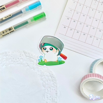 A sticker of Cappy from the cartoon Hamtaro standing on a patch of grass, photographed on a white desk background.