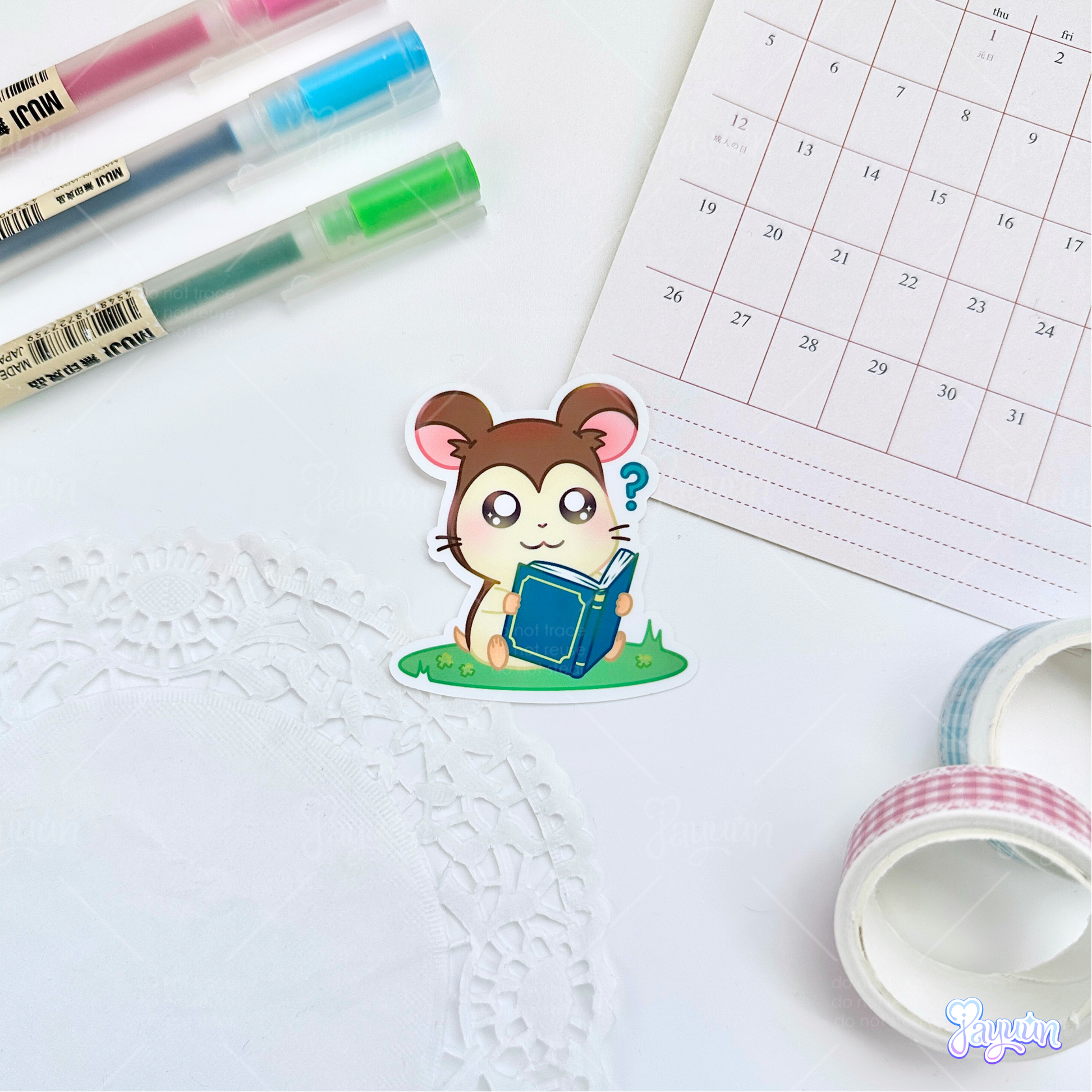 A sticker of Maxwell from the cartoon Hamtaro sitting on a patch of grass with a book, photographed on a white desk background.