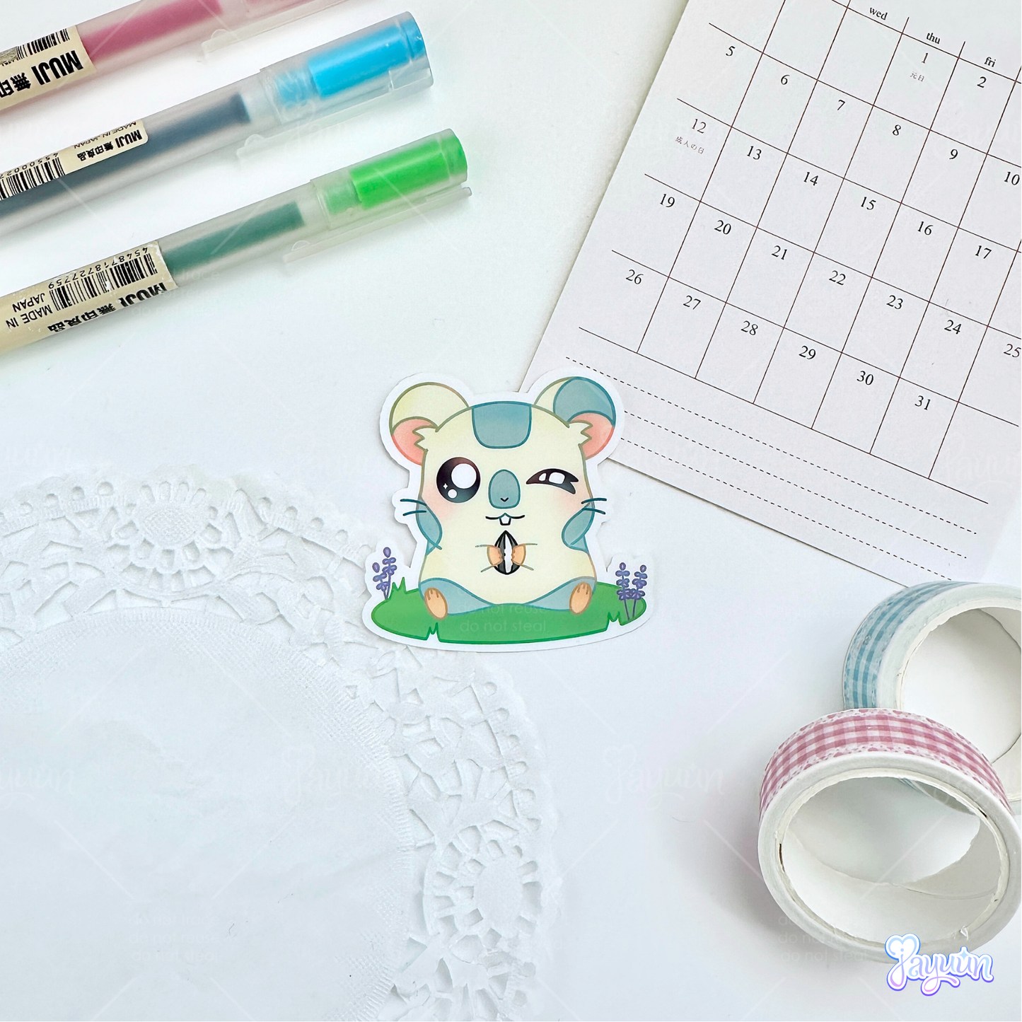 A sticker of Oxnard from the cartoon Hamtaro sitting on a patch of grass holding a sunflower seed, photographed on a white desk background.