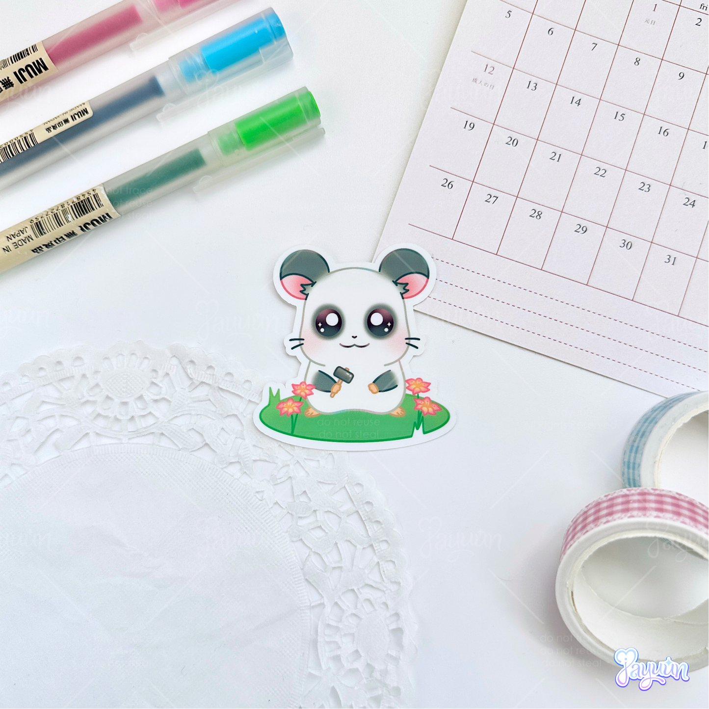 A sticker of Panda from the cartoon Hamtaro standing on a patch of grass holding a hammer, photographed on a white desk background.