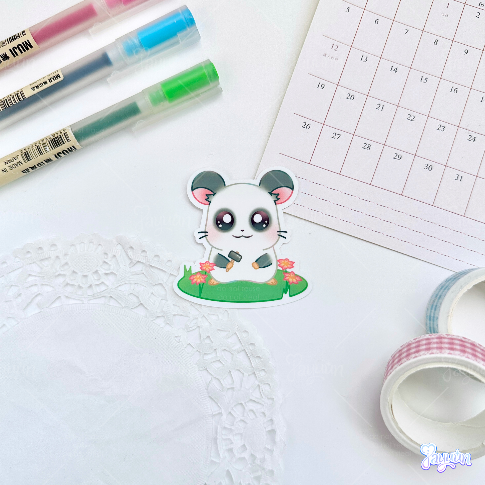 A sticker of Panda from the cartoon Hamtaro standing on a patch of grass holding a hammer, photographed on a white desk background.