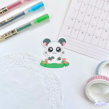 A sticker of Panda from the cartoon Hamtaro standing on a patch of grass holding a hammer, photographed on a white desk background.