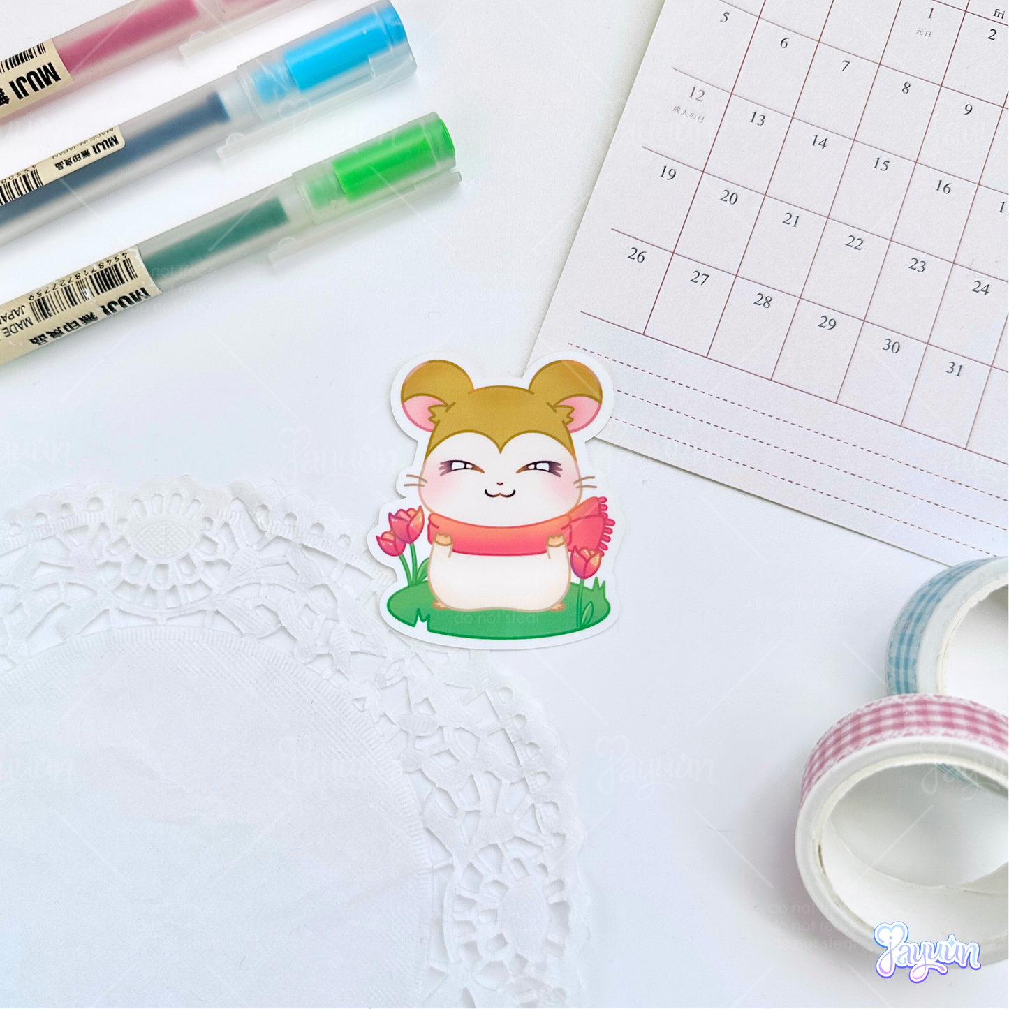 A sticker of Pashmina from the cartoon Hamtaro standing on a patch of grass, photographed on a white desk background.