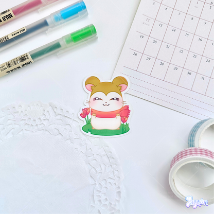 A sticker of Pashmina from the cartoon Hamtaro standing on a patch of grass, photographed on a white desk background.