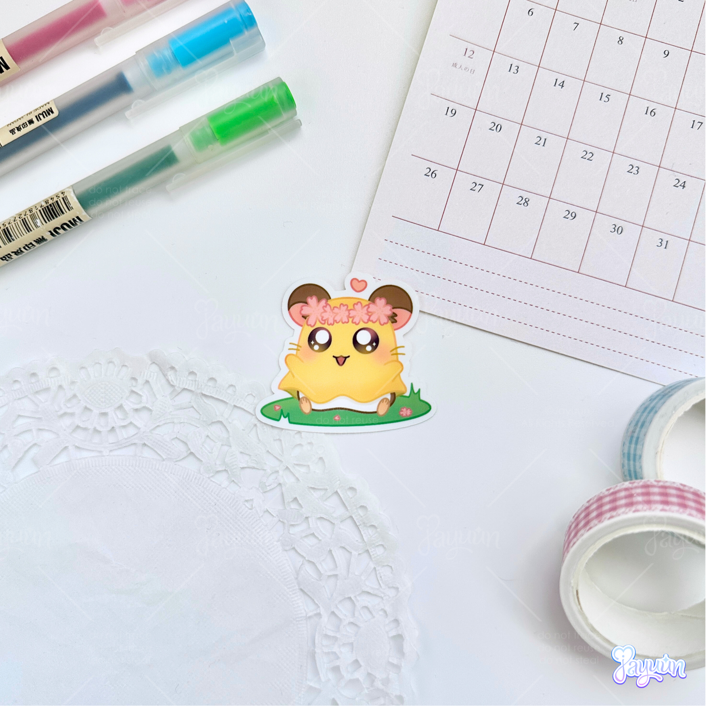 A sticker of Penelope from the cartoon Hamtaro sitting on a patch of grass, photographed on a white desk background.