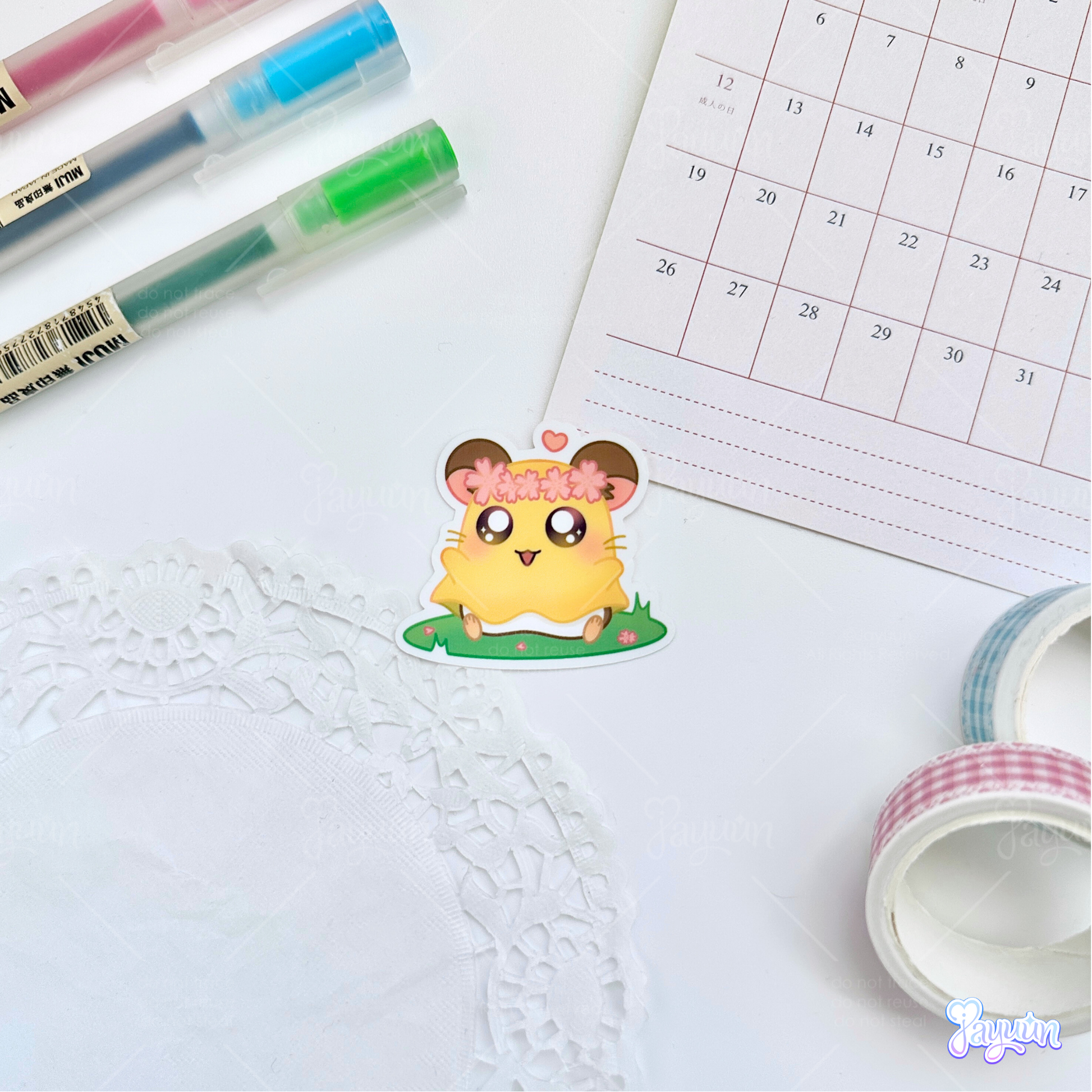 A sticker of Penelope from the cartoon Hamtaro sitting on a patch of grass, photographed on a white desk background.