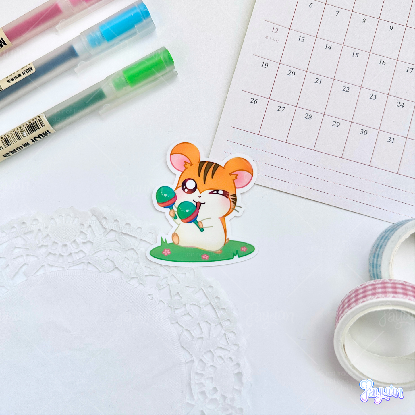 A sticker of Sam from the cartoon Hamtaro standing on a patch of grass with maracas, photographed on a white desk background.