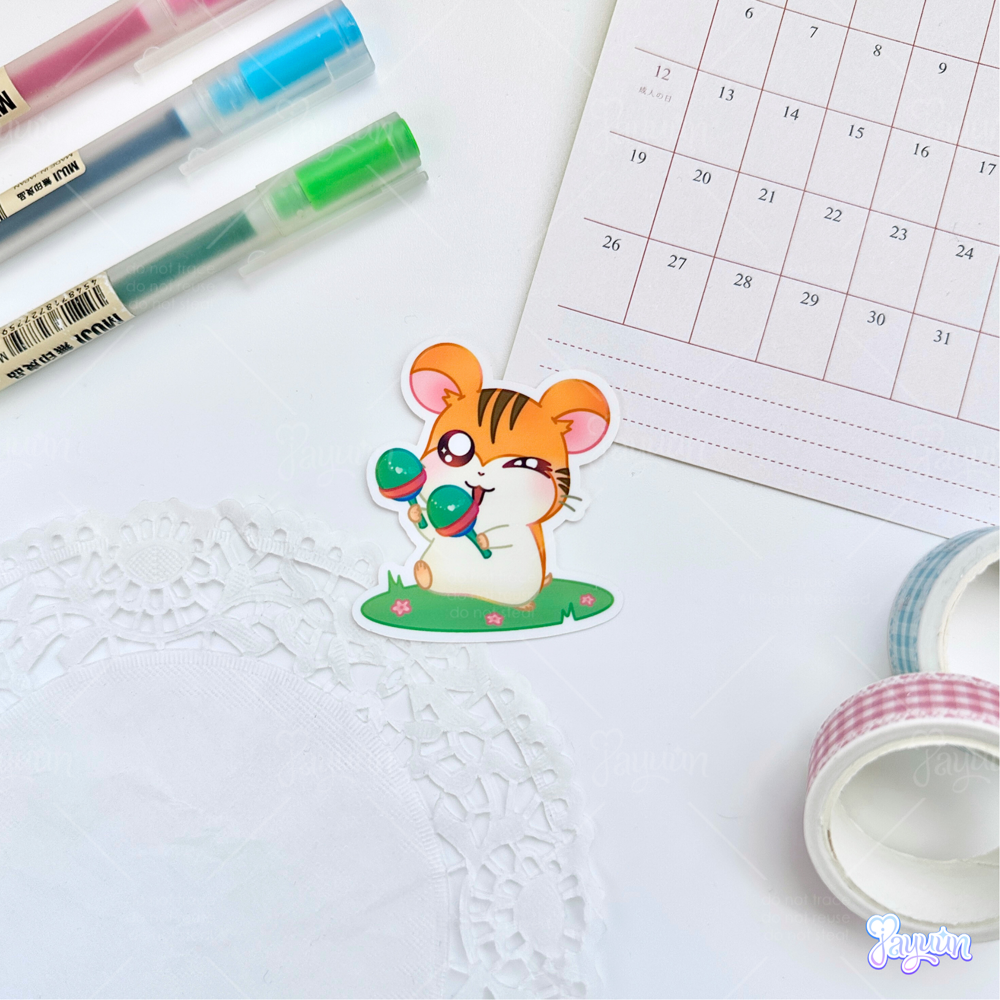 A sticker of Sam from the cartoon Hamtaro standing on a patch of grass with maracas, photographed on a white desk background.