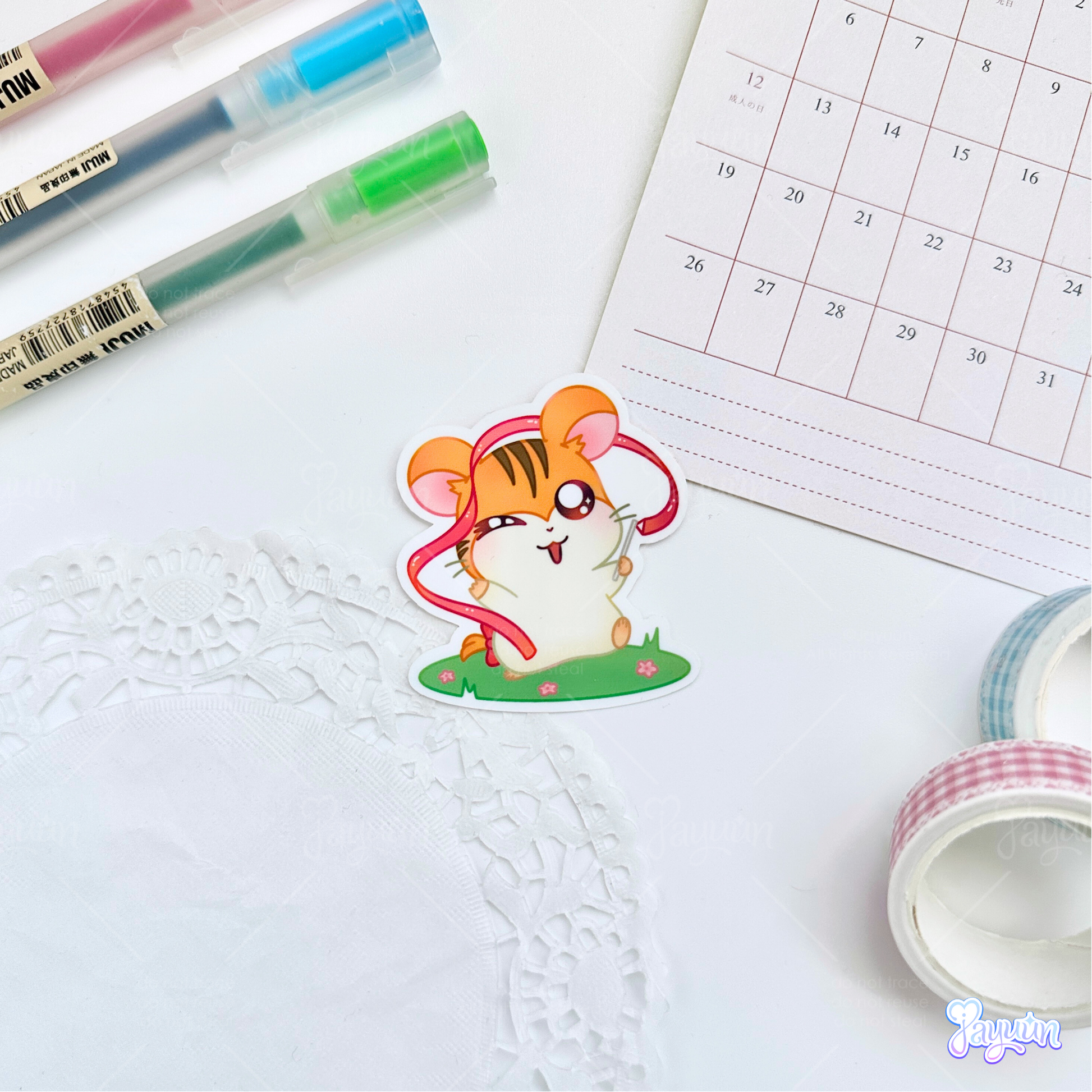 A sticker of Sandy from the cartoon Hamtaro standing on a patch of grass holding a ribbon, photographed on a white desk background.