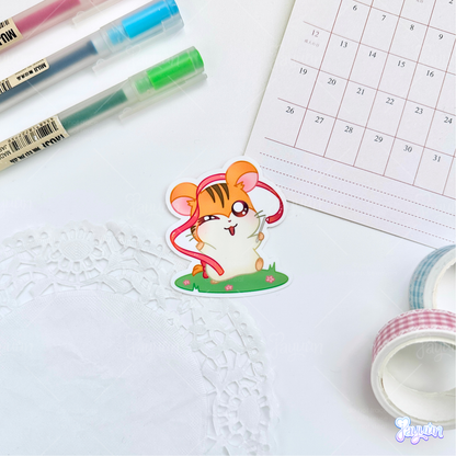 A sticker of Sandy from the cartoon Hamtaro standing on a patch of grass holding a ribbon, photographed on a white desk background.