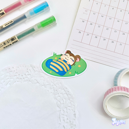 A sticker of Snoozer from the cartoon Hamtaro sleeping on a patch of grass , photographed on a white desk background.