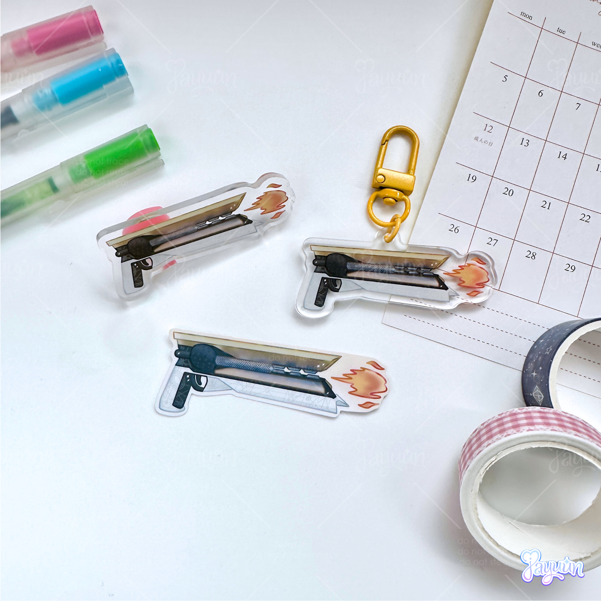 Solar hand cannon acrylic pin, acrylic charm, and sticker photographed on a white desk background.