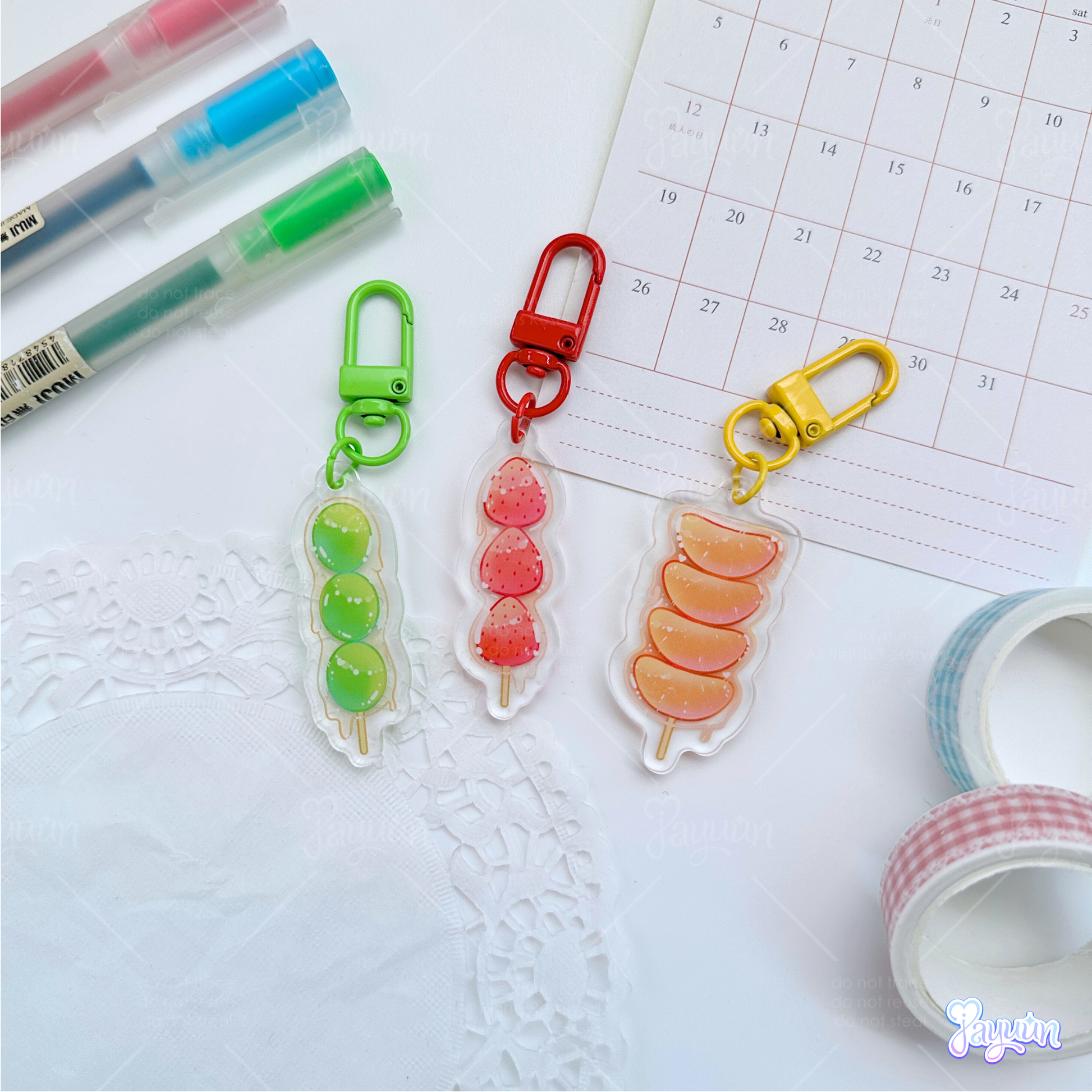 A green grape tanghulu acrylic charm, red strawberry tanghulu acrylic charm, and an orange tanghulu acrylic charm photographed on a white desk background.