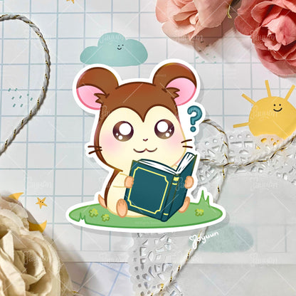 A sticker of Maxwell from the cartoon Hamtaro sitting on a patch of grass, shown on a decorative paper background,