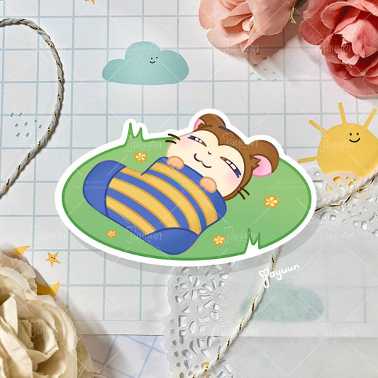 A sticker of Snoozer from the cartoon Hamtaro sleeping on a patch of grass, shown on a decorative paper background